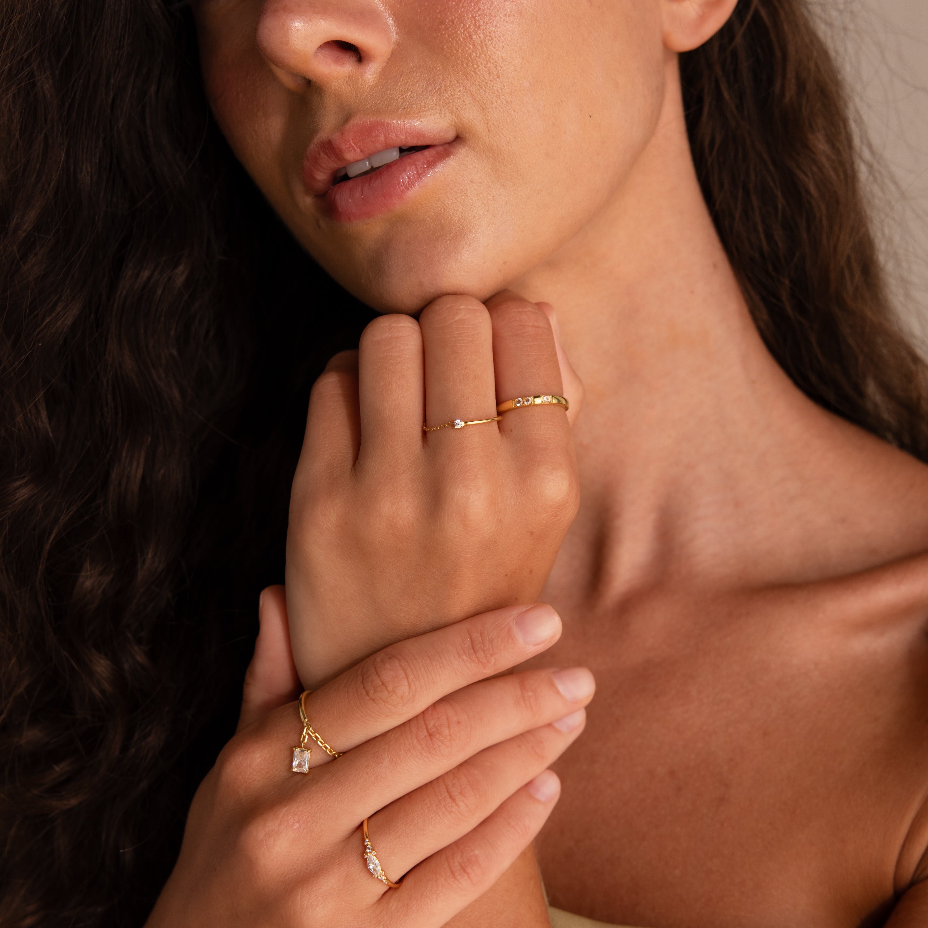 Woman with wavy hair wearing dainty jewelry, including a Diamond Half Chain Ring and a birthstone ring, gently touching her face and collarbone.