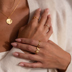Close-up of hands with a nude manicure, wearing the Hand & Footprint Duo Name Ring in Rose Gold as a personalized keepsake, plus gold rings and a gold pendant necklace, styled against a cream top.