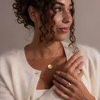 Woman with curly hair wears gold jewelry, including the Baby Hand & Footprint Necklace in Rose Gold, a cream cardigan, and looks to the side against a neutral background.