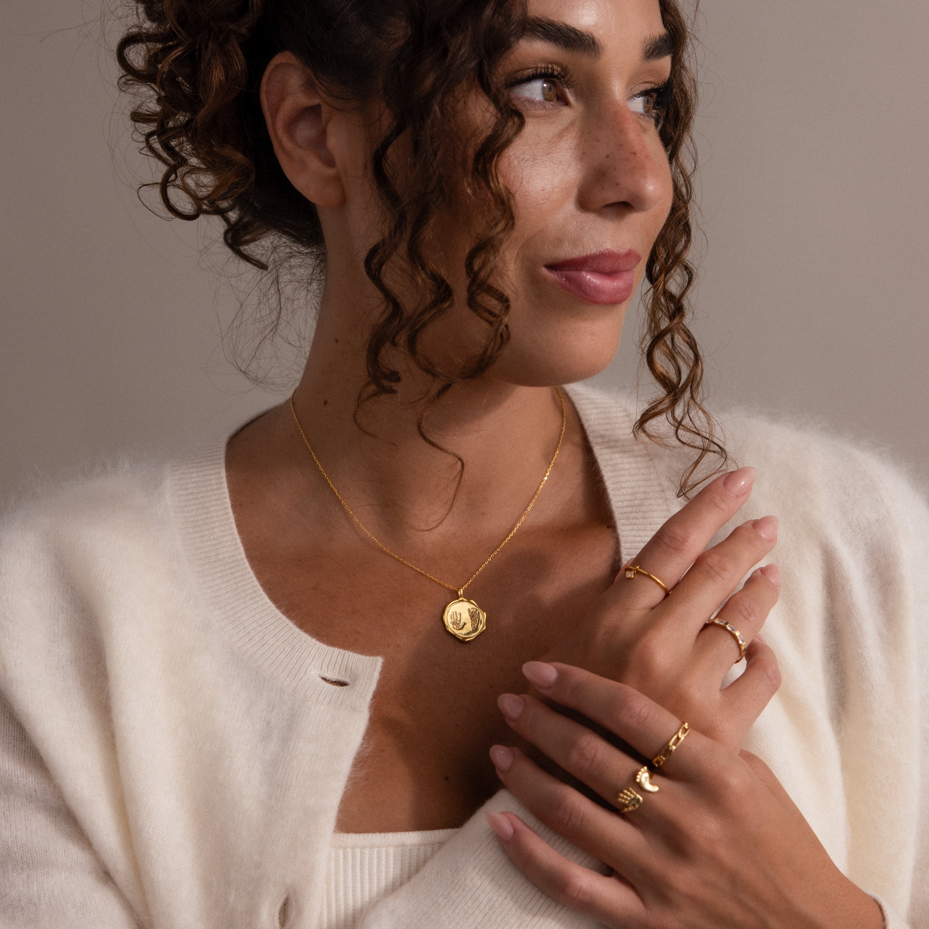 Woman with curly hair wears gold jewelry, including the Baby Hand & Footprint Necklace in Rose Gold, a cream cardigan, and looks to the side against a neutral background.