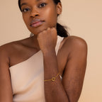 A woman in a cream one-shoulder top rests her chin on her hand, wearing the Rolo Circle Charm Bracelet in 18K Gold.