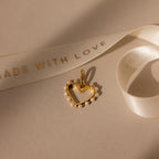 The Classic Pearl Heart Charm in 18K Gold, adorned with small pearls, is displayed beside a white ribbon printed with MADE WITH LOVE.