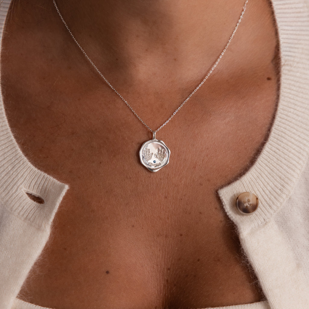 A woman wears the Baby Hand & Footprint Necklace in Sterling Silver with a cream cardigan, its button partially open.