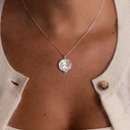A woman wears the Baby Hand & Footprint Necklace in Sterling Silver with a cream cardigan, its button partially open.