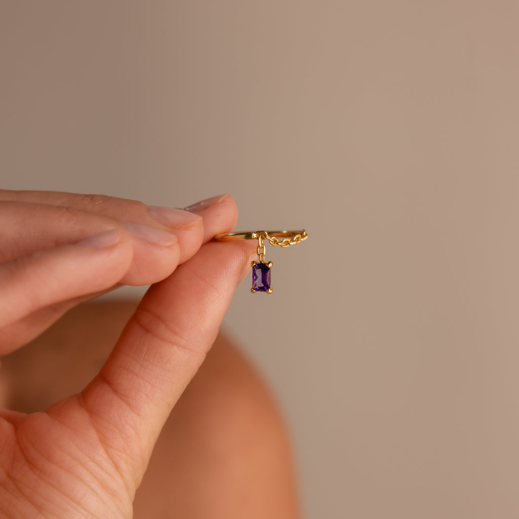 A hand holds the Birthstone Drop Chain Ring featuring a small dangling purple gemstone charm against a neutral background.