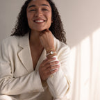 A smiling woman in a cream blazer rests her chin on her hand, highlighting bracelets and the Pearl Link Timepiece with diamond accents.
