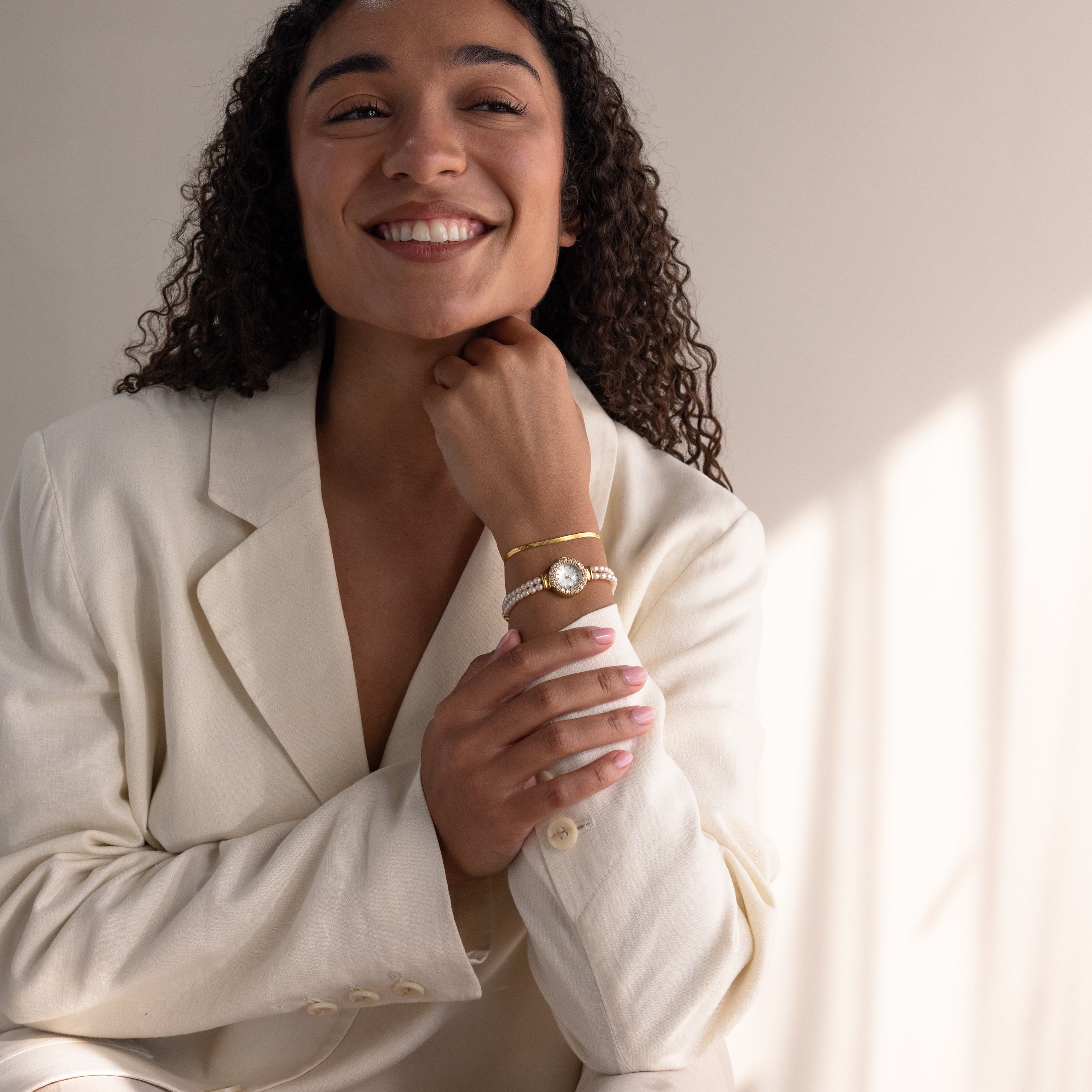 A smiling woman in a cream blazer rests her chin on her hand, highlighting bracelets and the Pearl Link Timepiece with diamond accents.