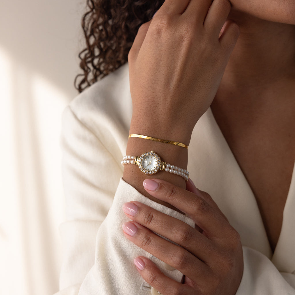 Woman in a cream blazer wears a gold bracelet, neatly manicured nails, and the Pearl Link Timepiece with diamond accents.