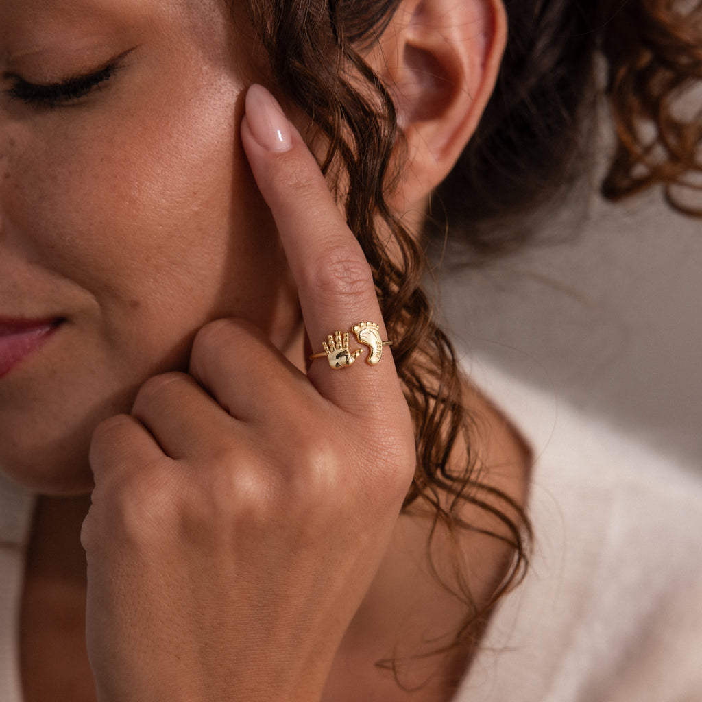 A woman touches her face, wearing the Hand & Footprint Duo Name Ring in 18K Gold—an ideal keepsake gift for new parents who treasure meaningful jewelry. Close-up shot.