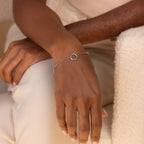 A hand wearing the Rolo Circle Charm Bracelet in White Gold, featuring a detachable hinged circle charm, rests on a cream surface—ideal for anyone who loves to personalize with charms.