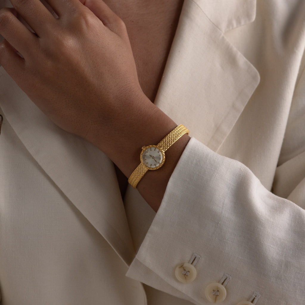 Close-up of a person wearing a gold round braided watch with a mesh strap, styled over a cream blazer.