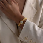Close-up of a person wearing a gold round braided watch with a mesh strap, styled over a cream blazer.