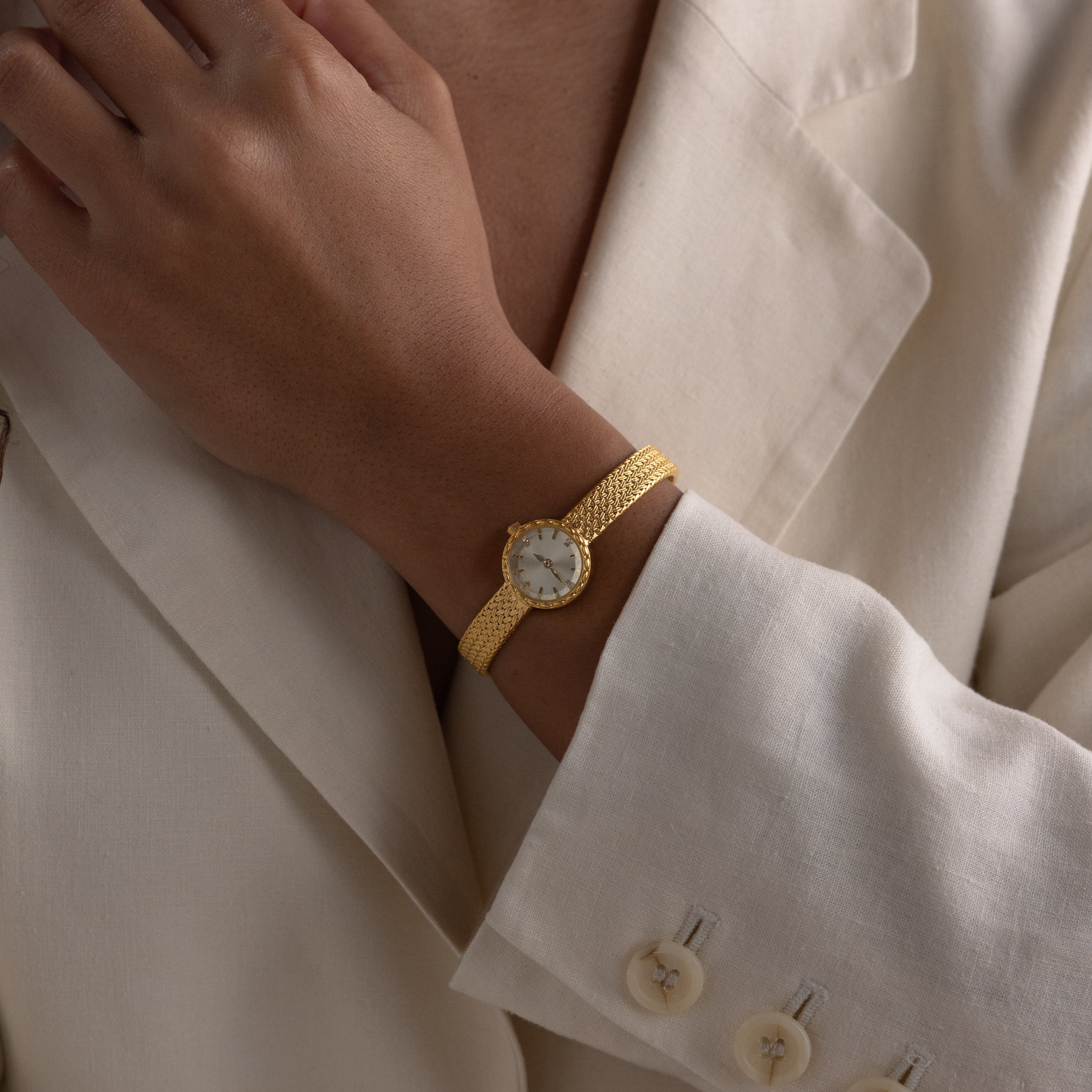 Close-up of a person wearing a gold round braided watch with a mesh strap, styled over a cream blazer.