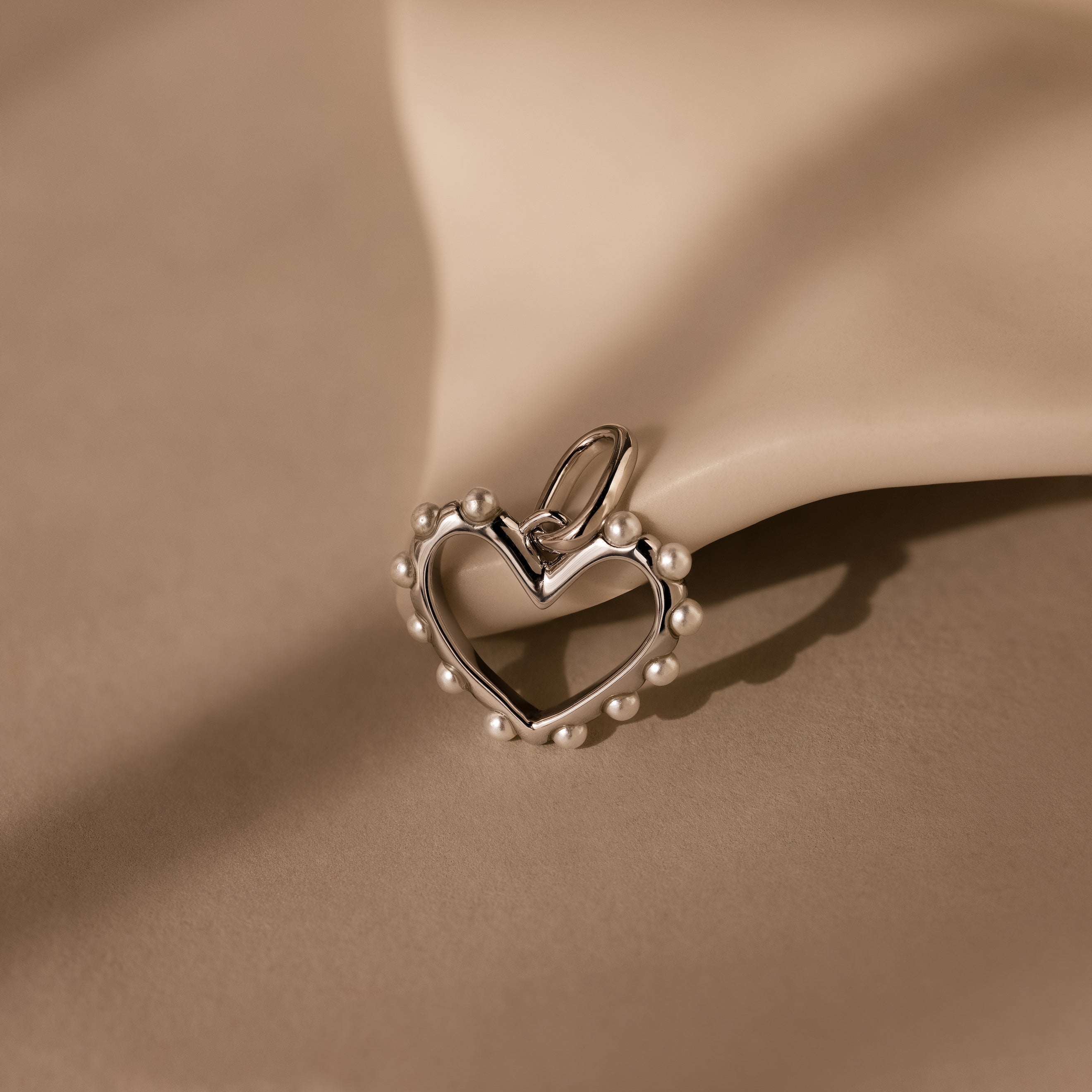 A Classic Pearl Heart Charm in Sterling Silver, adorned with pearl bead accents, rests on a smooth beige surface, evoking timeless elegance.