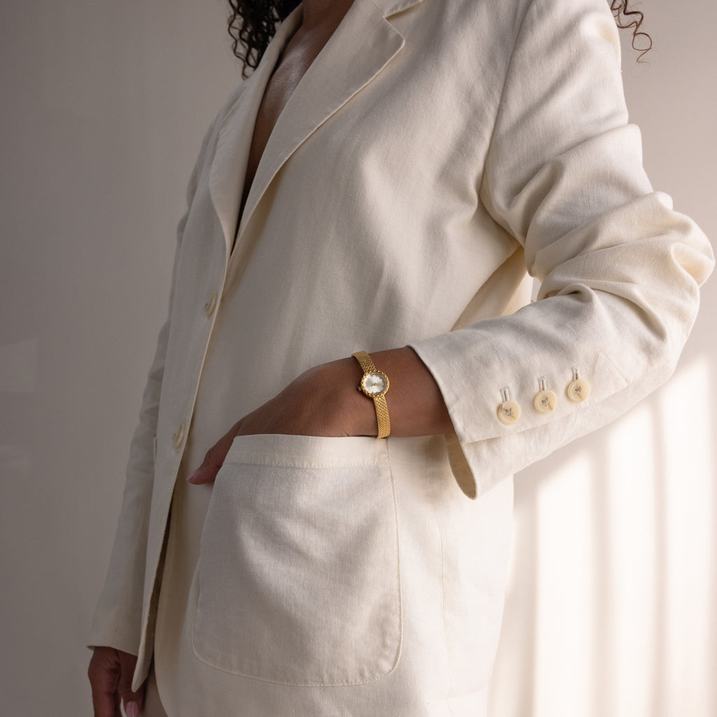 Side view of the gold watch worn on a woman’s wrist, one hand in the pocket of a matching cream suit.