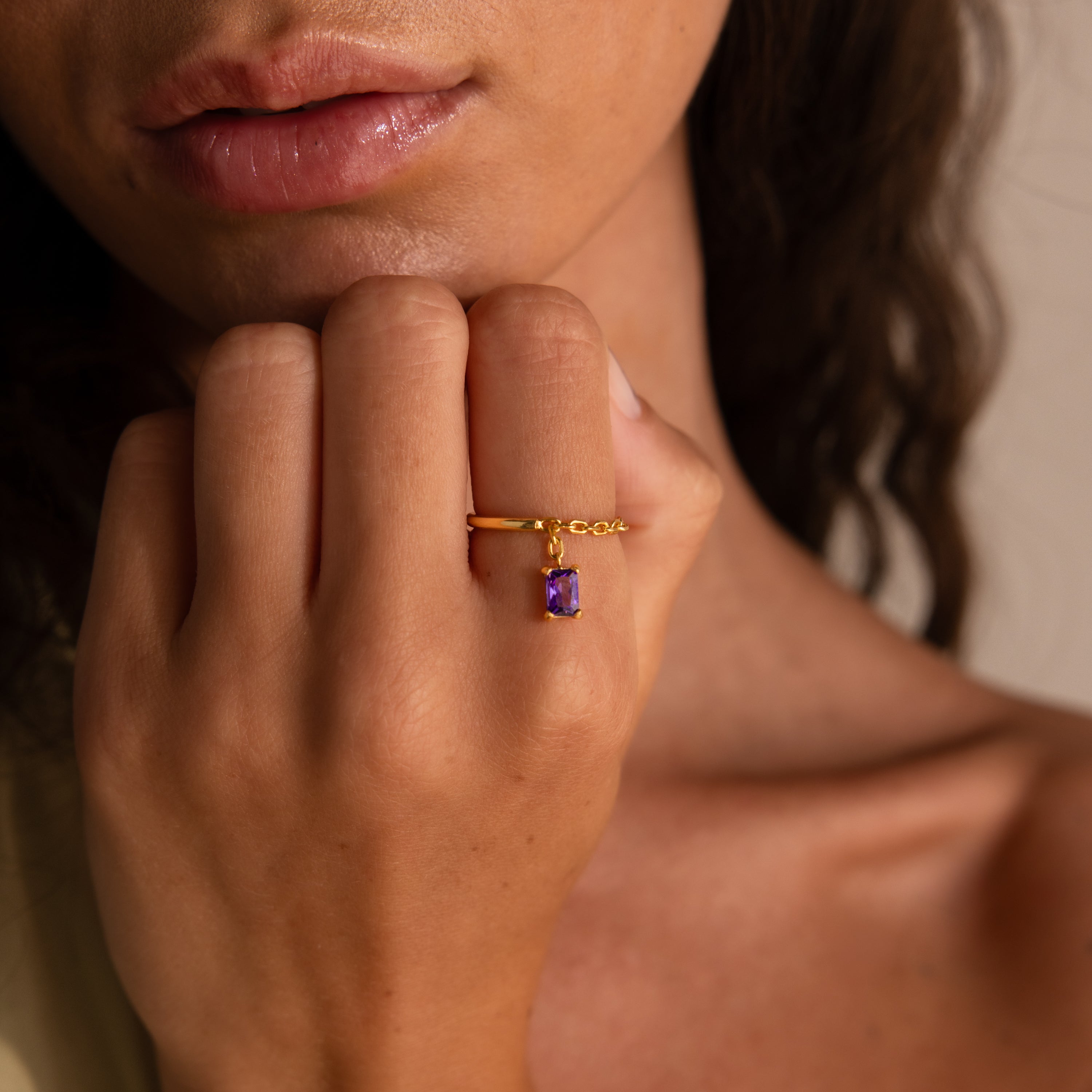 A woman with wavy hair wears the Birthstone Drop Chain Ring, featuring an emerald-cut purple gemstone, and rests her hand on her chin.
