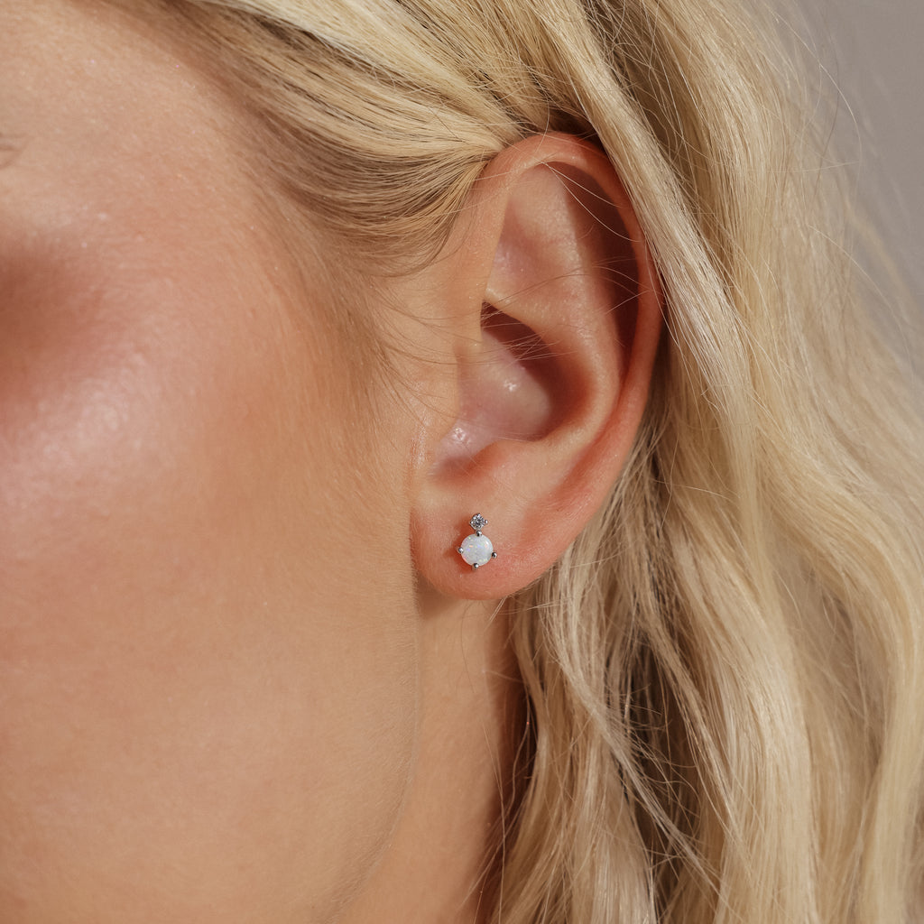 Close-up of a woman's ear wearing a silver flatback stud earring featuring a round opal stone with a tiny diamond above it.