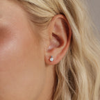 Close-up of a woman's ear wearing a silver flatback stud earring featuring a round opal stone with a tiny diamond above it.
