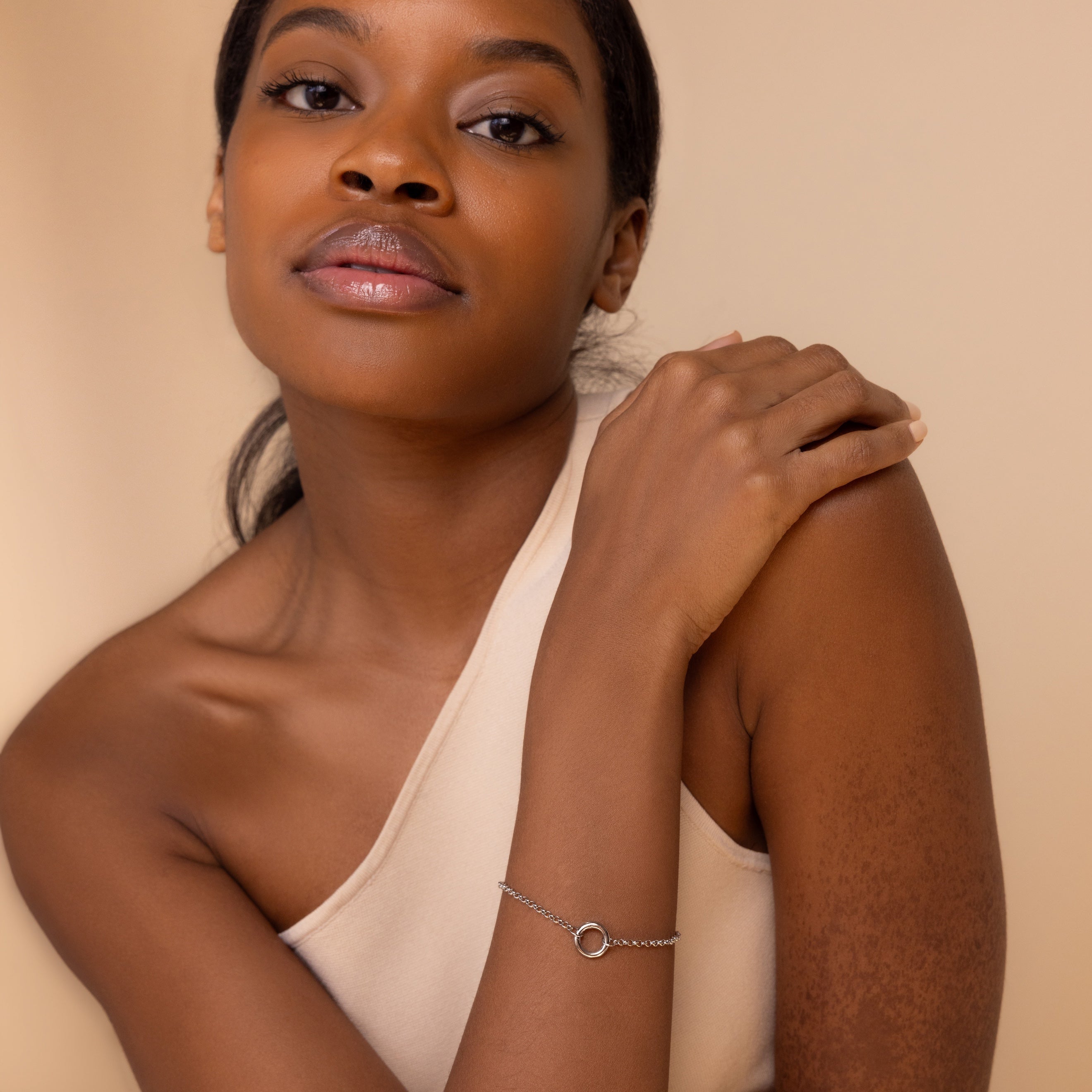 Woman in a one-shoulder top wears the Rolo Circle Charm Bracelet in White Gold, featuring a detachable hinged circle for adding charms, as she poses with her hand on her shoulder against a beige background.
