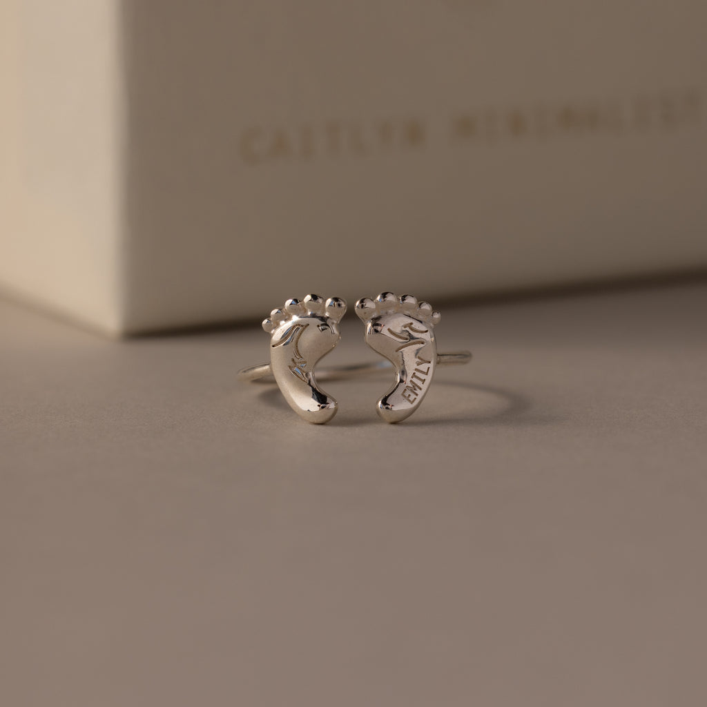 The Hand & Footprint Duo Name Ring in Sterling Silver features two baby foot charms, one engraved with EMILY—a perfect keepsake gift for new parents.