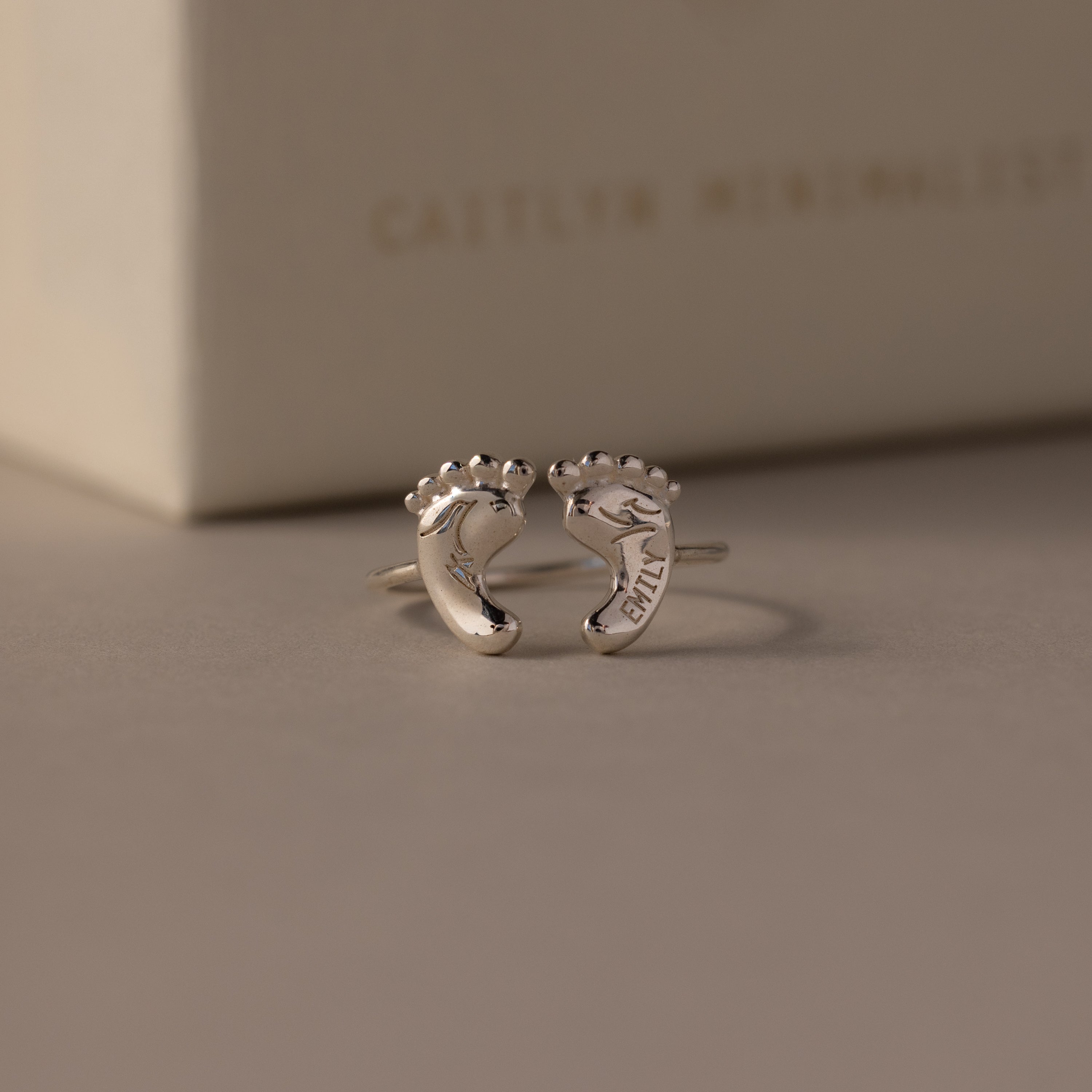 The Hand & Footprint Duo Name Ring features two baby feet with engraved names, making it a perfect personalized gift for new parents. Presented before a cream-colored box, this keepsake jewelry will be cherished forever.