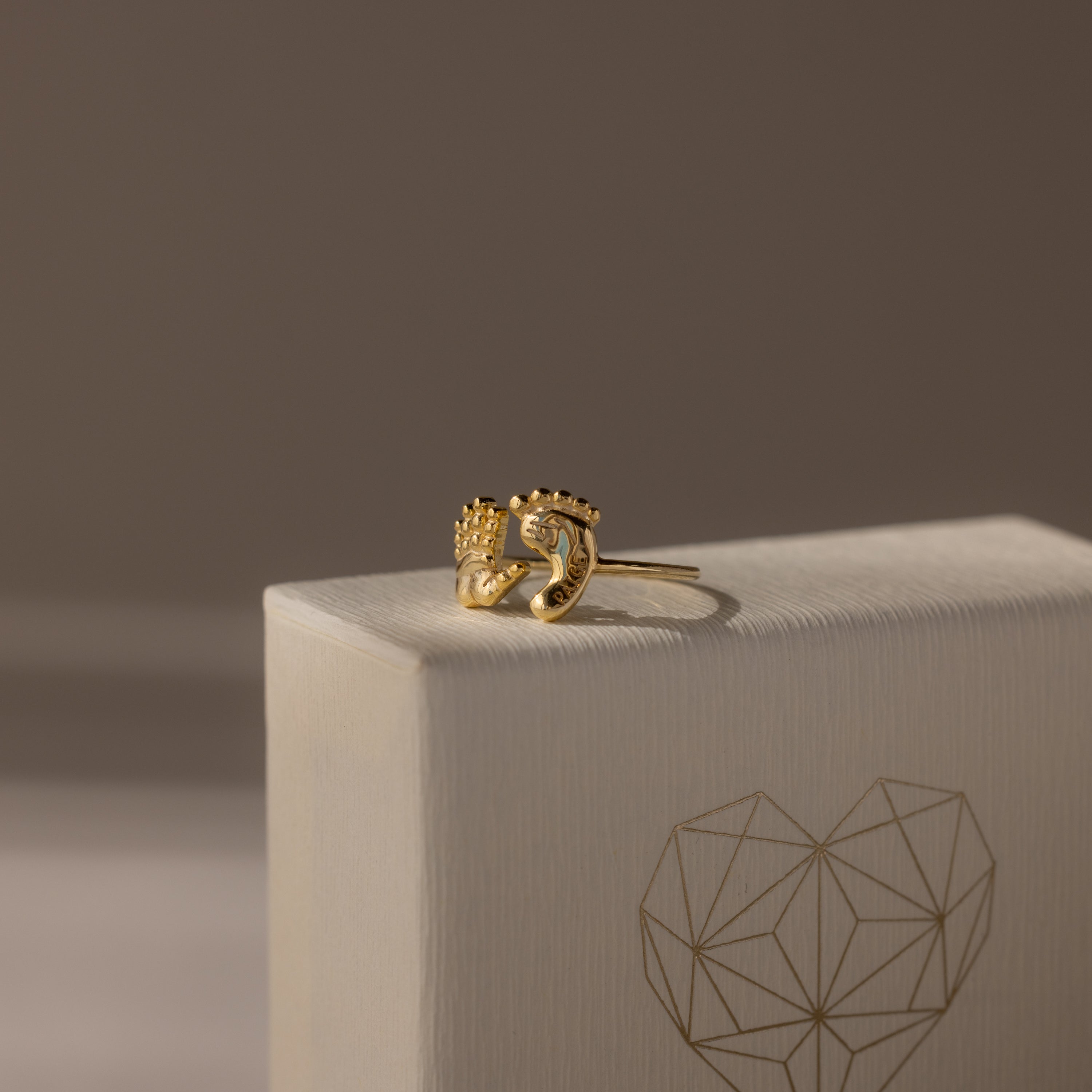 The Hand & Footprint Duo Name Ring in 18K Gold features a unique hand and foot design, displayed on a cream box with a geometric heart pattern—an ideal keepsake gift for new parents.