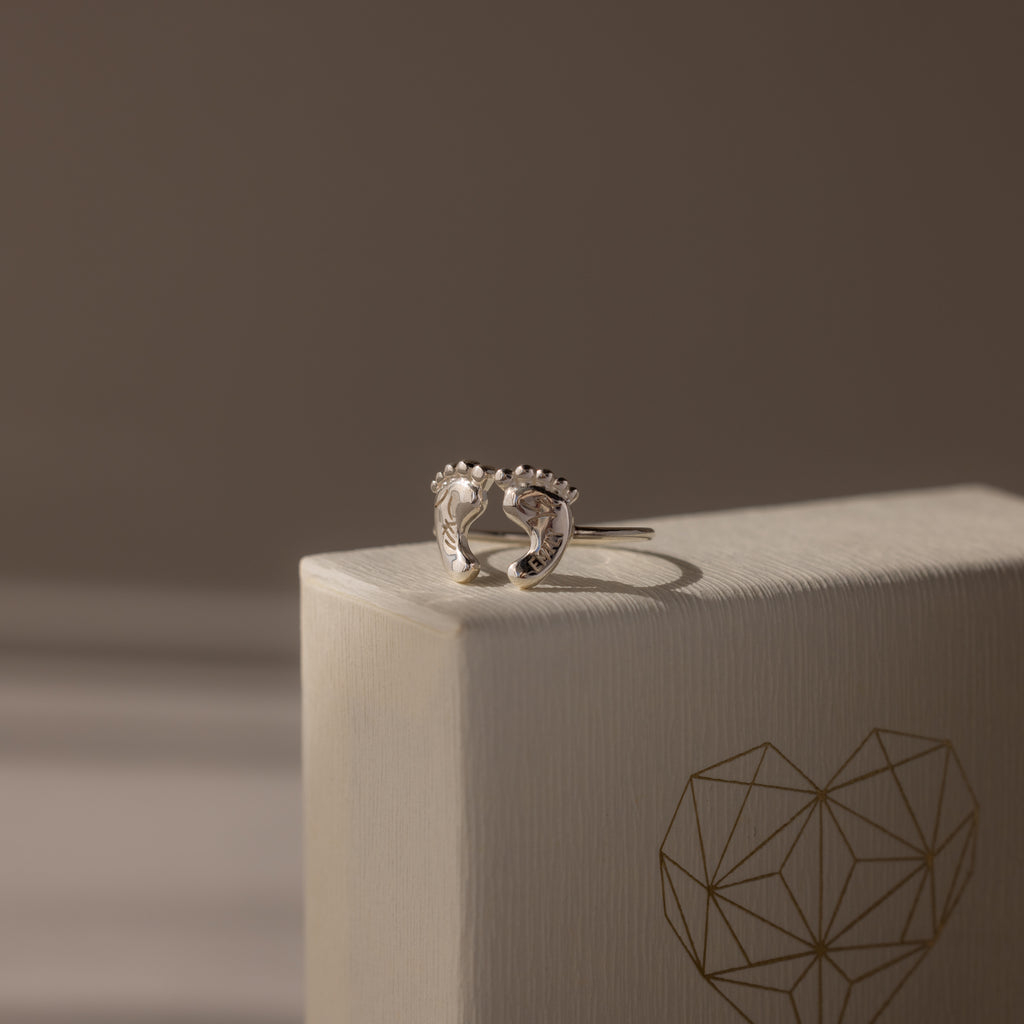 Hand & Footprint Duo Name Ring in Sterling Silver, a perfect keepsake gift for new parents. Presented on a textured box with a geometric heart illustration.