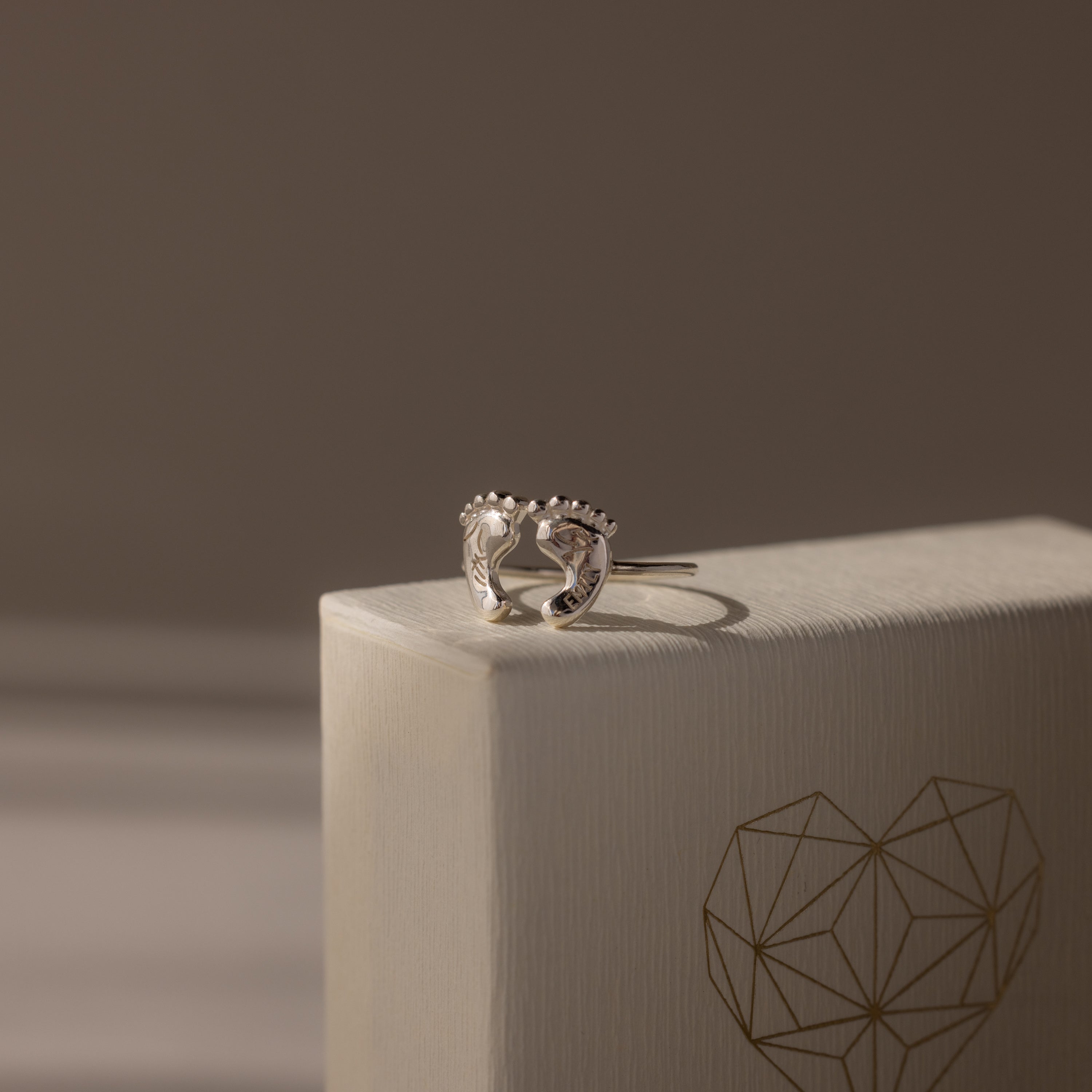 Hand & Footprint Duo Name Ring in Sterling Silver, a perfect keepsake gift for new parents. Presented on a textured box with a geometric heart illustration.