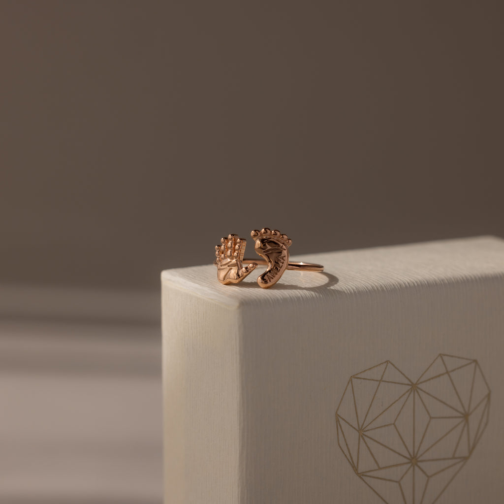 The Hand & Footprint Duo Name Ring in Rose Gold, featuring delicate hand and foot charms on a textured geometric heart box, makes a perfect personalized keepsake jewelry gift for new parents.