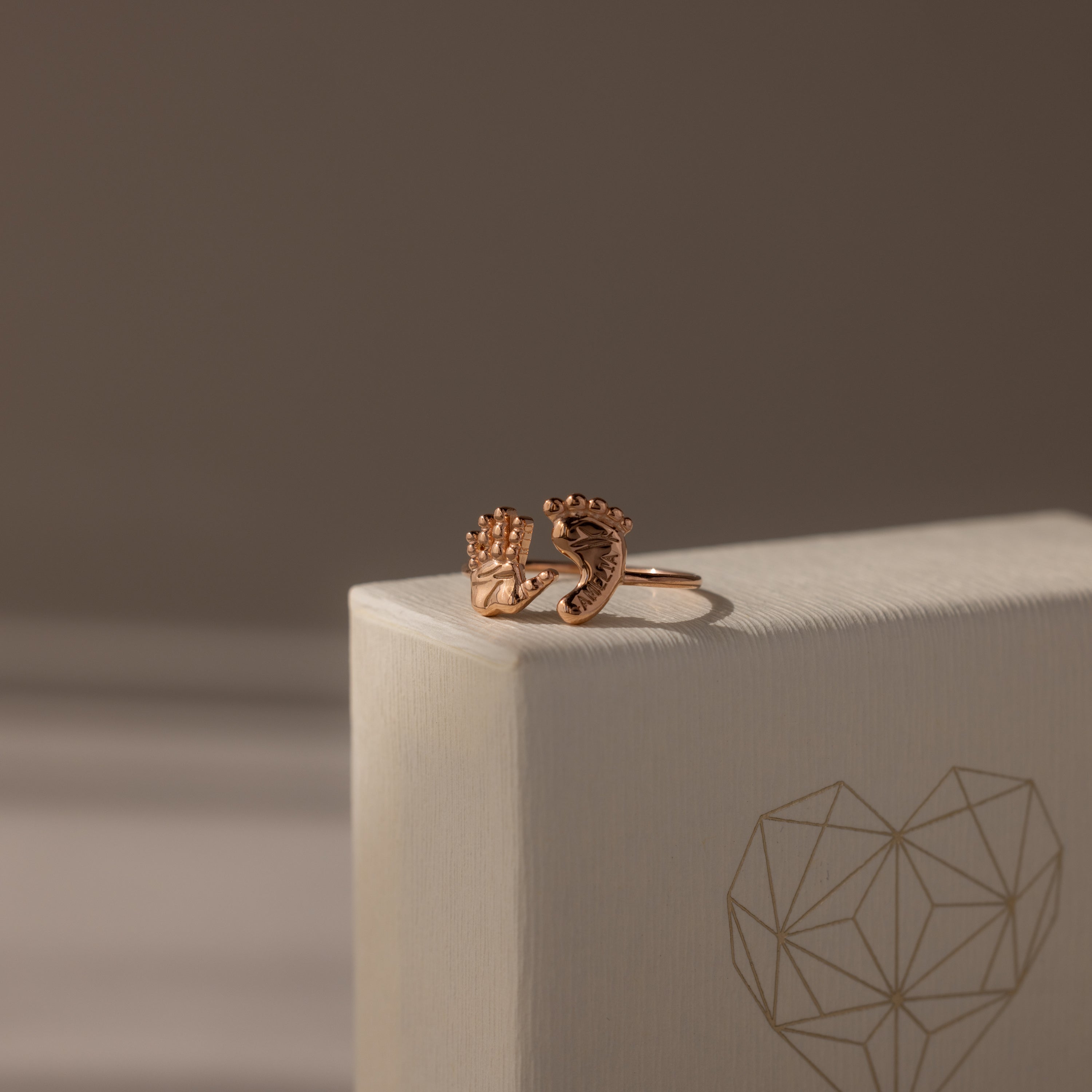 The Hand & Footprint Duo Name Ring in Rose Gold, featuring delicate hand and foot charms on a textured geometric heart box, makes a perfect personalized keepsake jewelry gift for new parents.