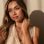 Woman with long hair in a cream ruffled top touches her Abalone Sunrise Pendant Necklace in White Gold, adorned with abalone and pearl gemstones, as she gazes thoughtfully to the side.