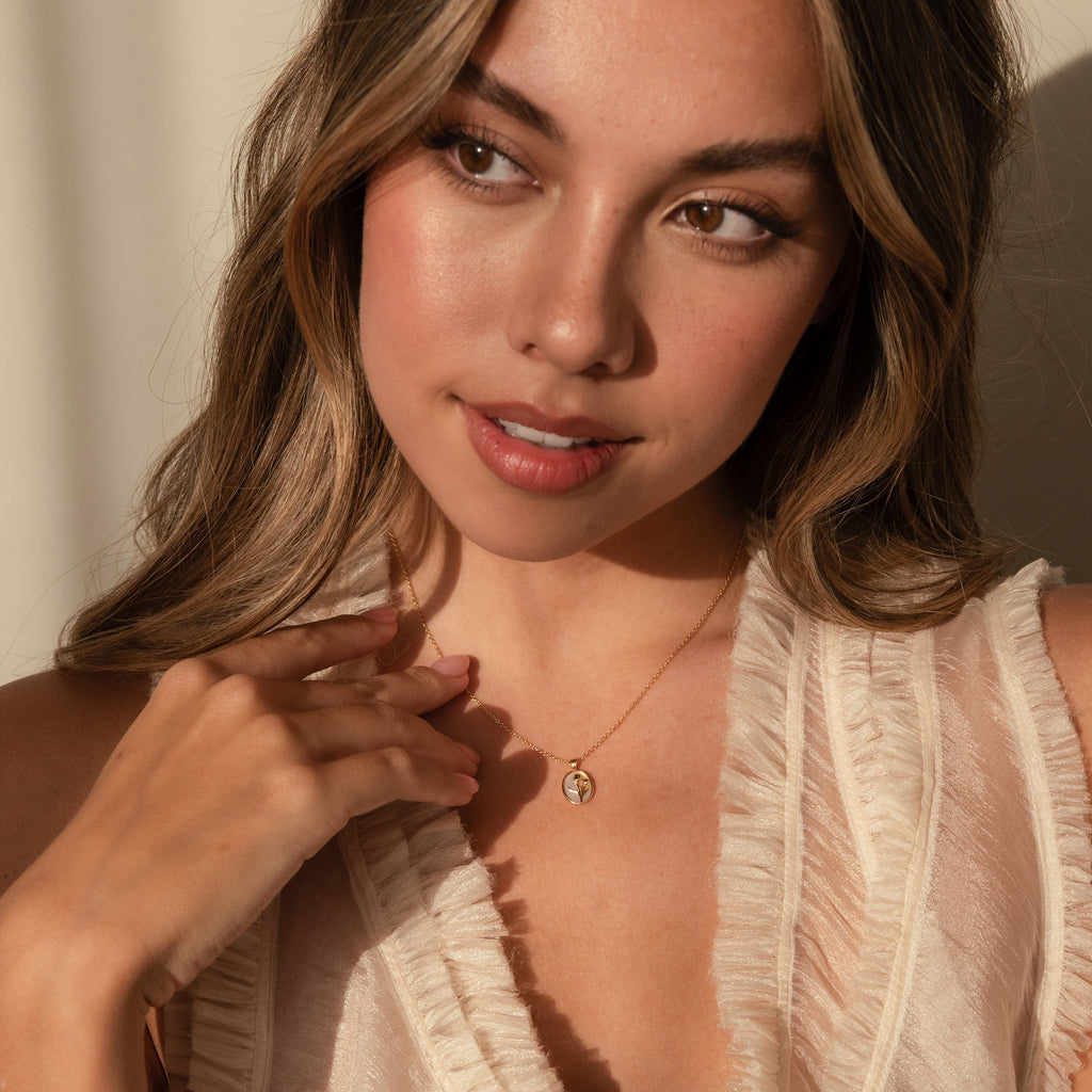 A woman with wavy hair, wearing a cream ruffled top in soft lighting, touches a Pearl Tulip Necklace in 18K Gold.