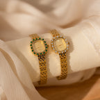 Two elegant Pave Blossom Timepiece watches—one with green CZ gemstones, the other with clear—rest on soft fabric. These jeweled bezels add a touch of luxury to any look.