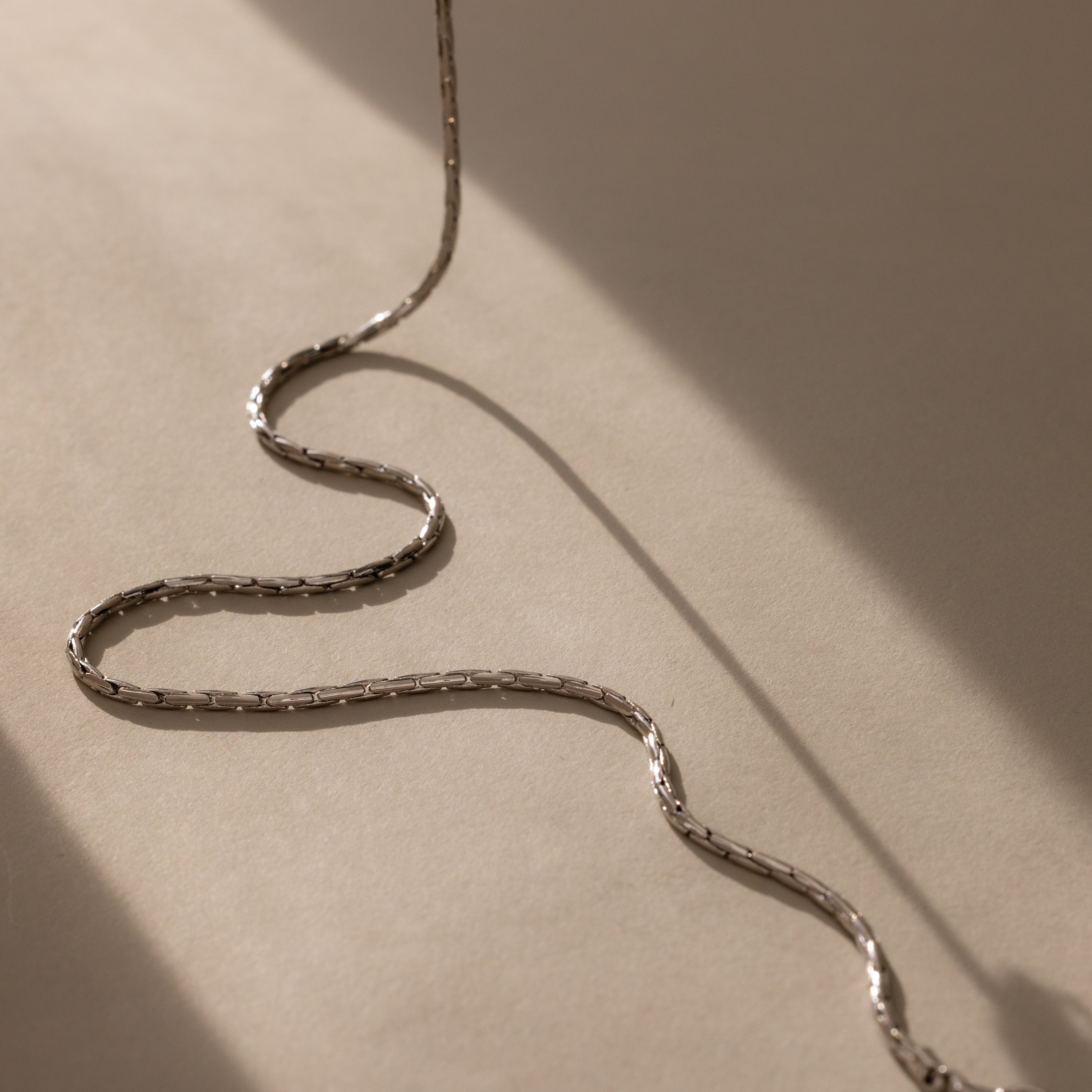 The Bold Link Chain Necklace in Silver rests in a wavy line on a beige surface, highlighted by soft shadows.