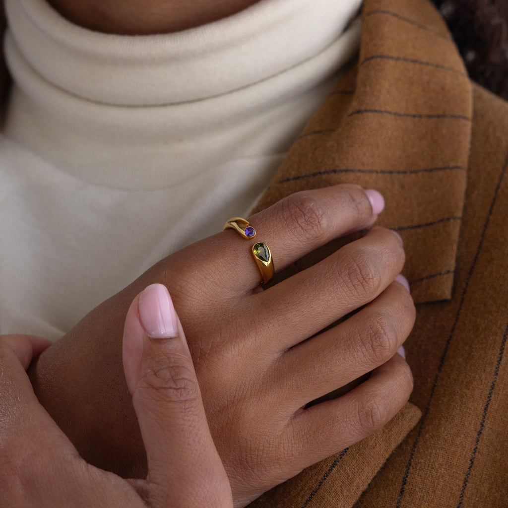 A hand with pink nails wears a gold Toi et Moi Birthstone Signet Ring featuring two gemstones, resting on a brown pinstripe blazer.