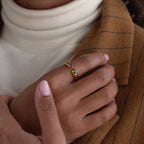 A hand with pink nails wears a gold Toi et Moi Birthstone Signet Ring featuring two gemstones, resting on a brown pinstripe blazer.