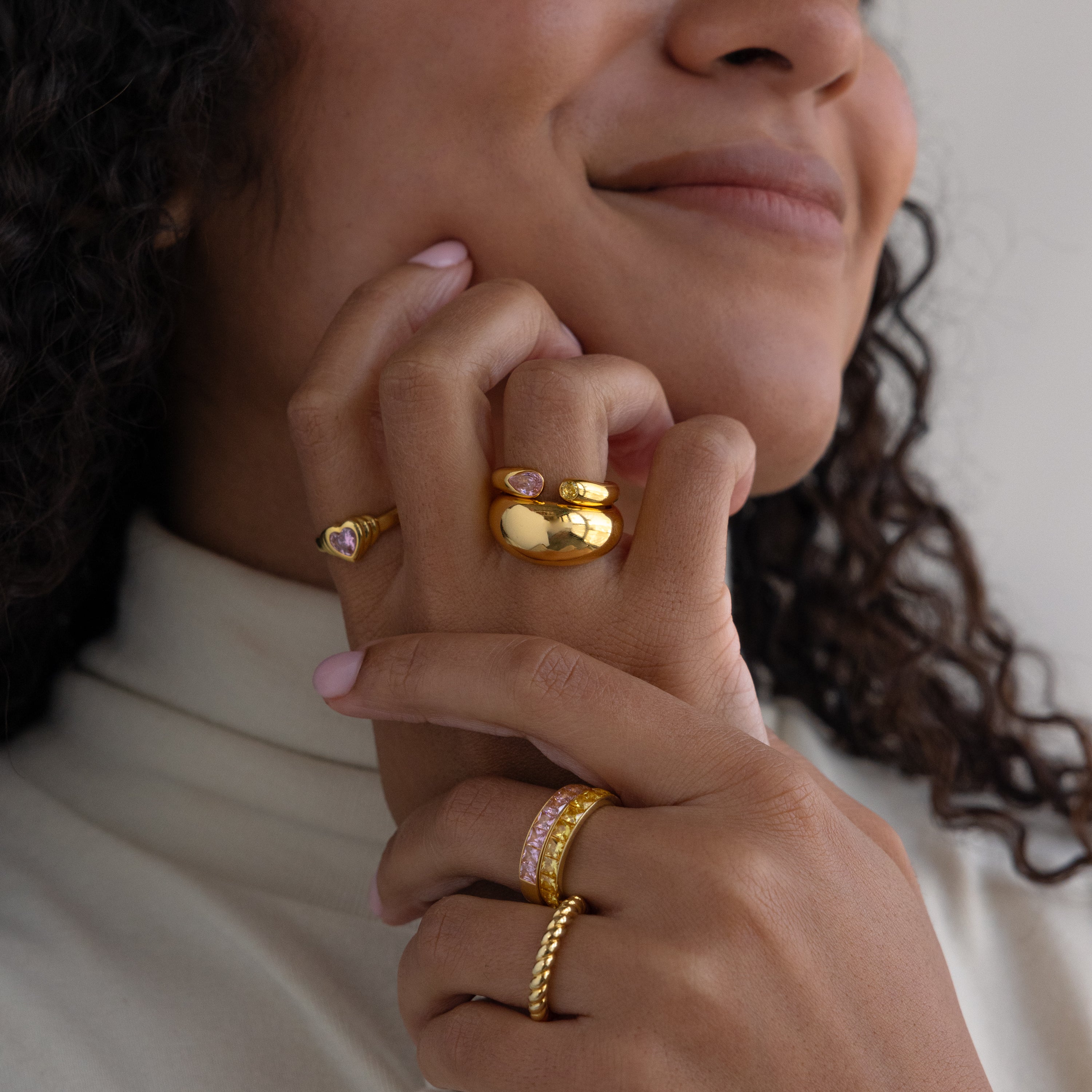 A woman with curly hair and a cream top smiles, displaying her Toi et Moi Birthstone Signet Ring and gold bands on her fingers.