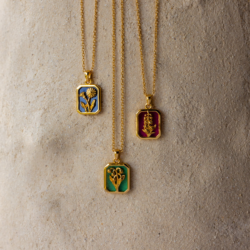 The Ruby Birth Flower Charm Necklace features a gold floral pendant with delicate ruby red enamel, resting elegantly on a stone surface and capturing timeless beauty.