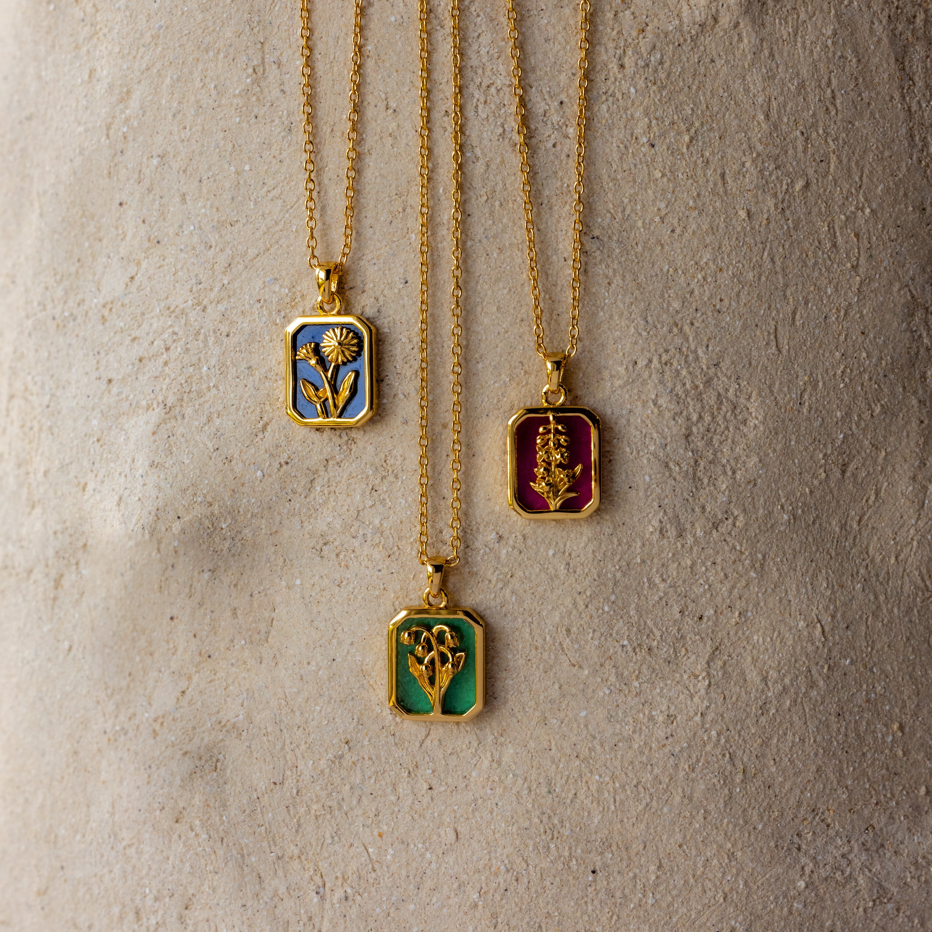 The Birth Flower Charm Necklace features a gold chain with a vibrant enamel floral pendant, displayed against a textured beige background—the perfect artistic touch for your jewelry collection.