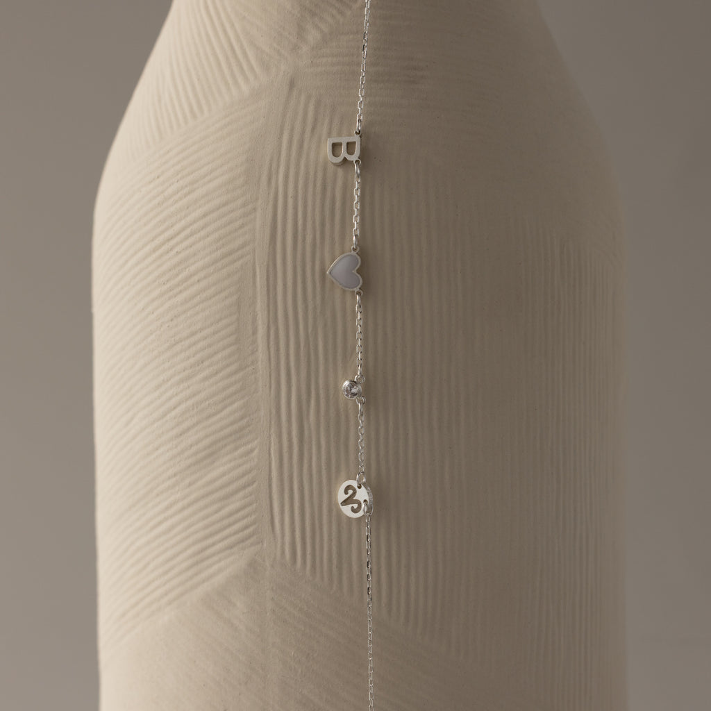 The Dainty Space Charm Necklace in Sterling Silver, featuring letter, heart, bead, and number charms, drapes beautifully—ideal to pair with a silver chain bracelet for a unique personalized style.