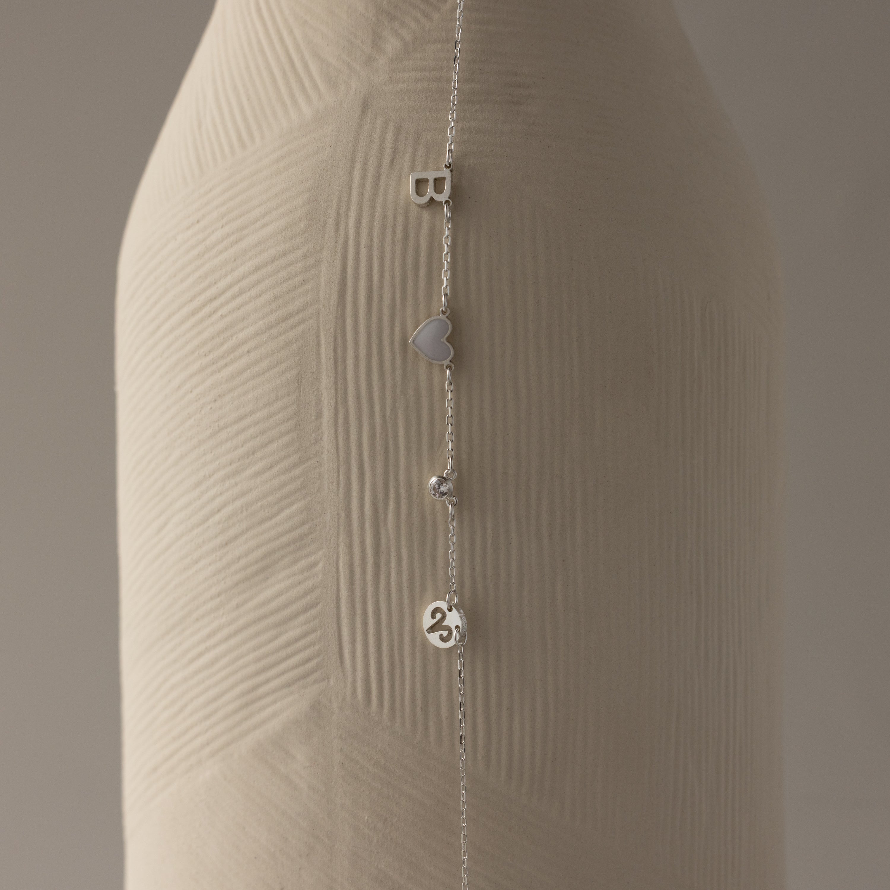 The Dainty Space Charm Necklace in Sterling Silver, featuring letter, heart, bead, and number charms, drapes beautifully—ideal to pair with a silver chain bracelet for a unique personalized style.