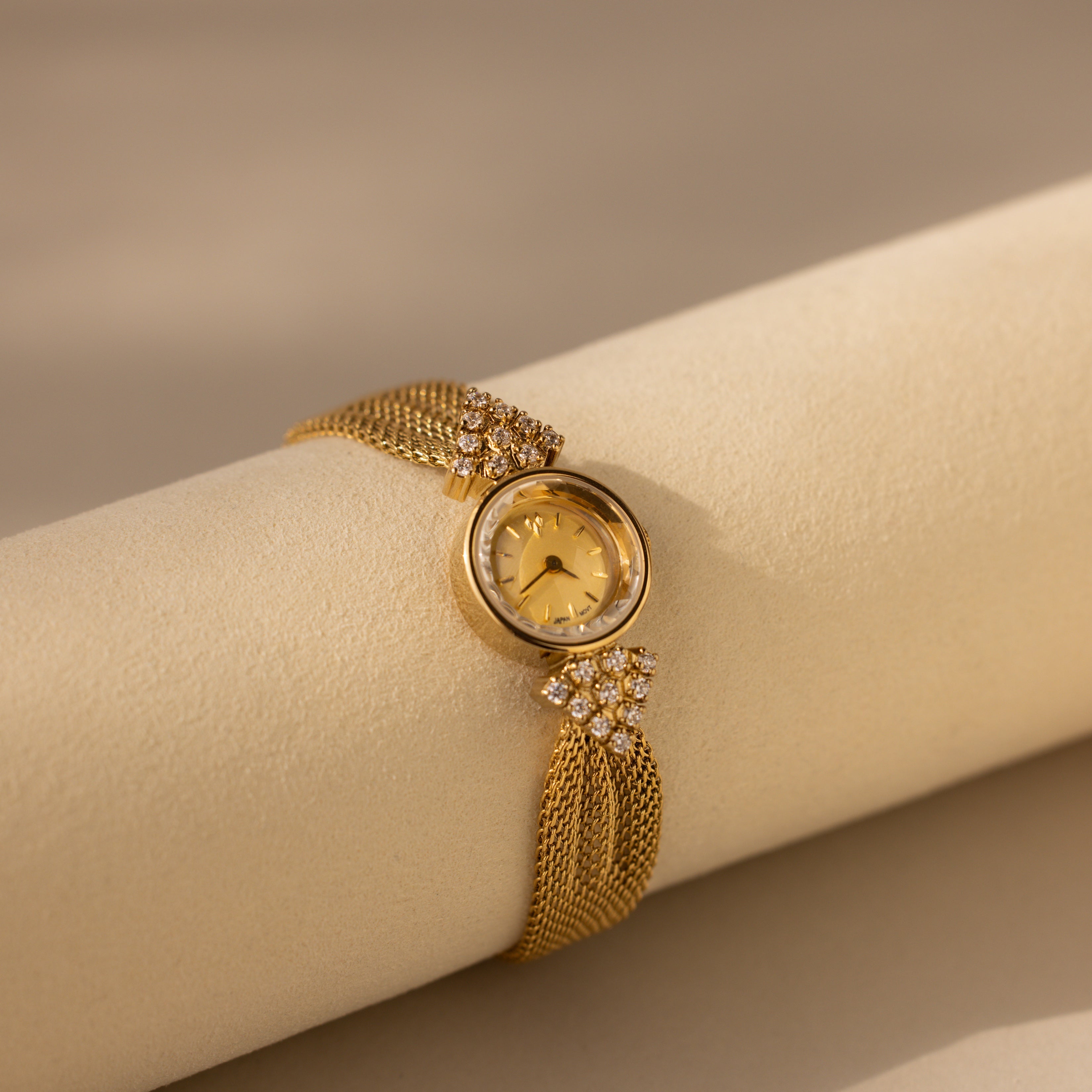The Diamond Mesh Timepiece features a gold mesh band and diamond accents, offering vintage elegance as it rests on a soft beige surface.