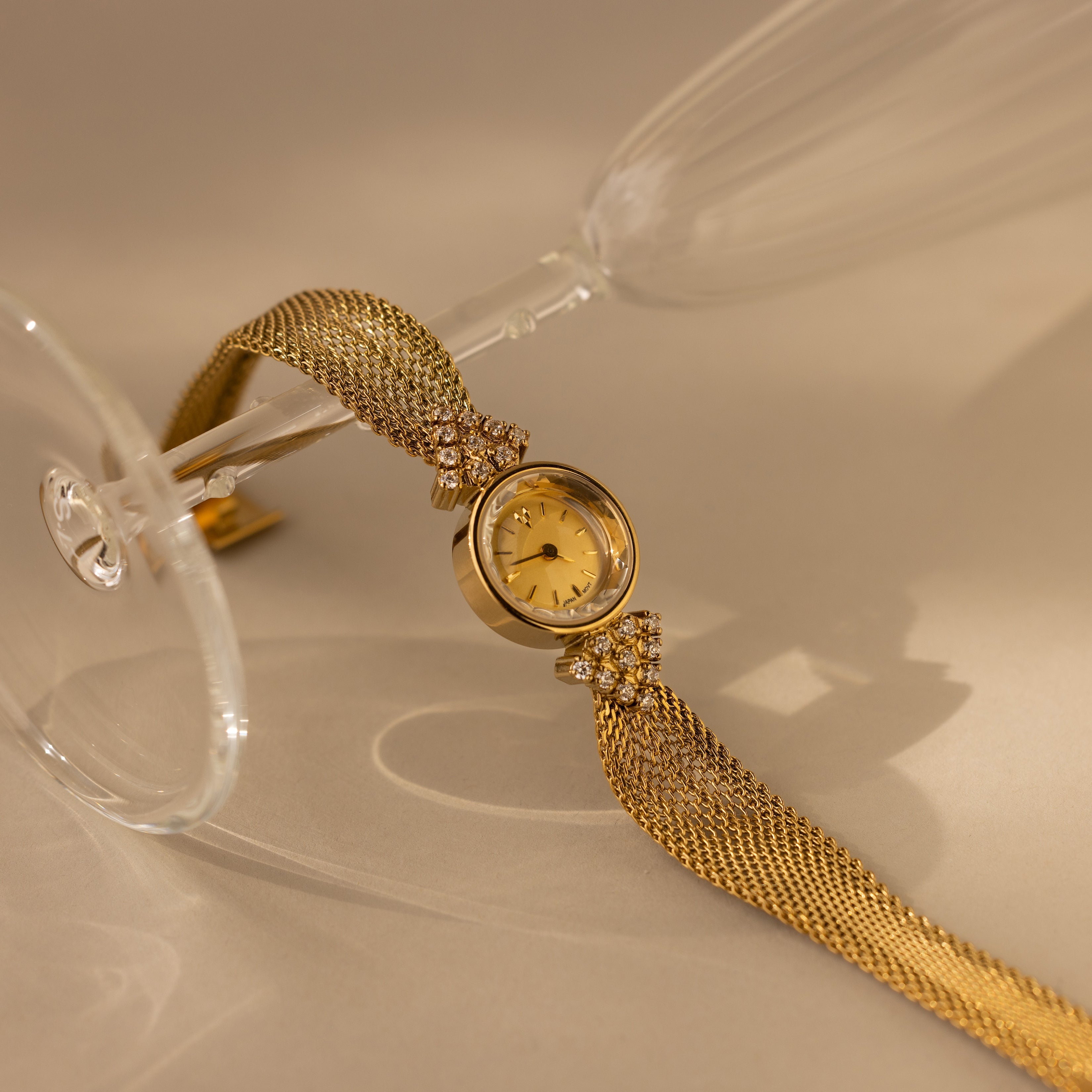 The Diamond Mesh Timepiece features a gold mesh band and is elegantly displayed on a clear wine glass against a beige background.