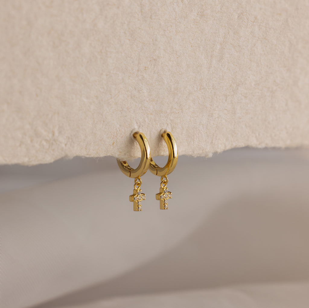 Pave Cross Huggies—gold dainty hoops with dangling cross charms—are displayed on a textured beige surface.