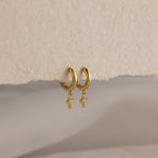 Pave Cross Huggies—gold dainty hoops with dangling cross charms—are displayed on a textured beige surface.