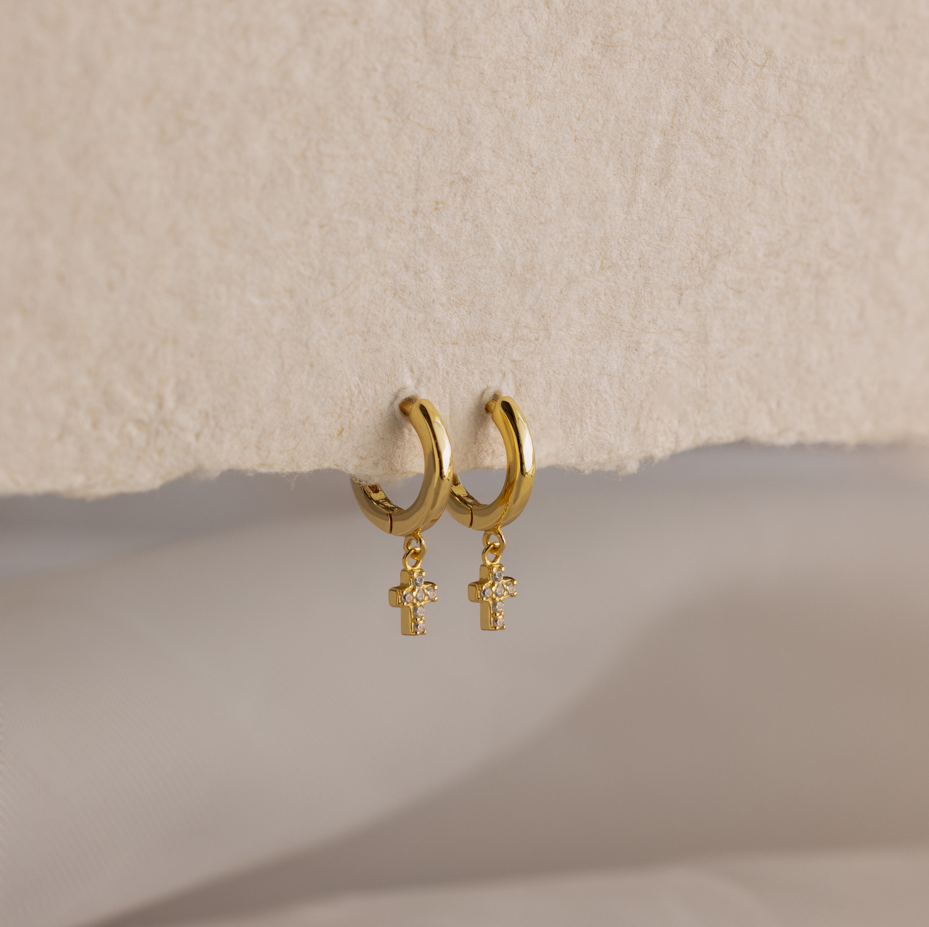 Pave Cross Huggies—gold dainty hoops with dangling cross charms—are displayed on a textured beige surface.