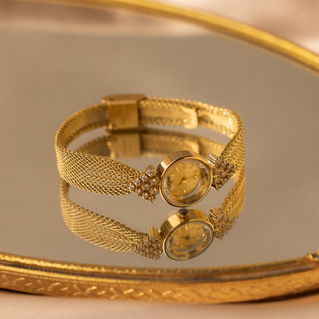 The Diamond Mesh Timepiece features a gold mesh band, round face, and diamond accents, shown on a reflective gold tray.