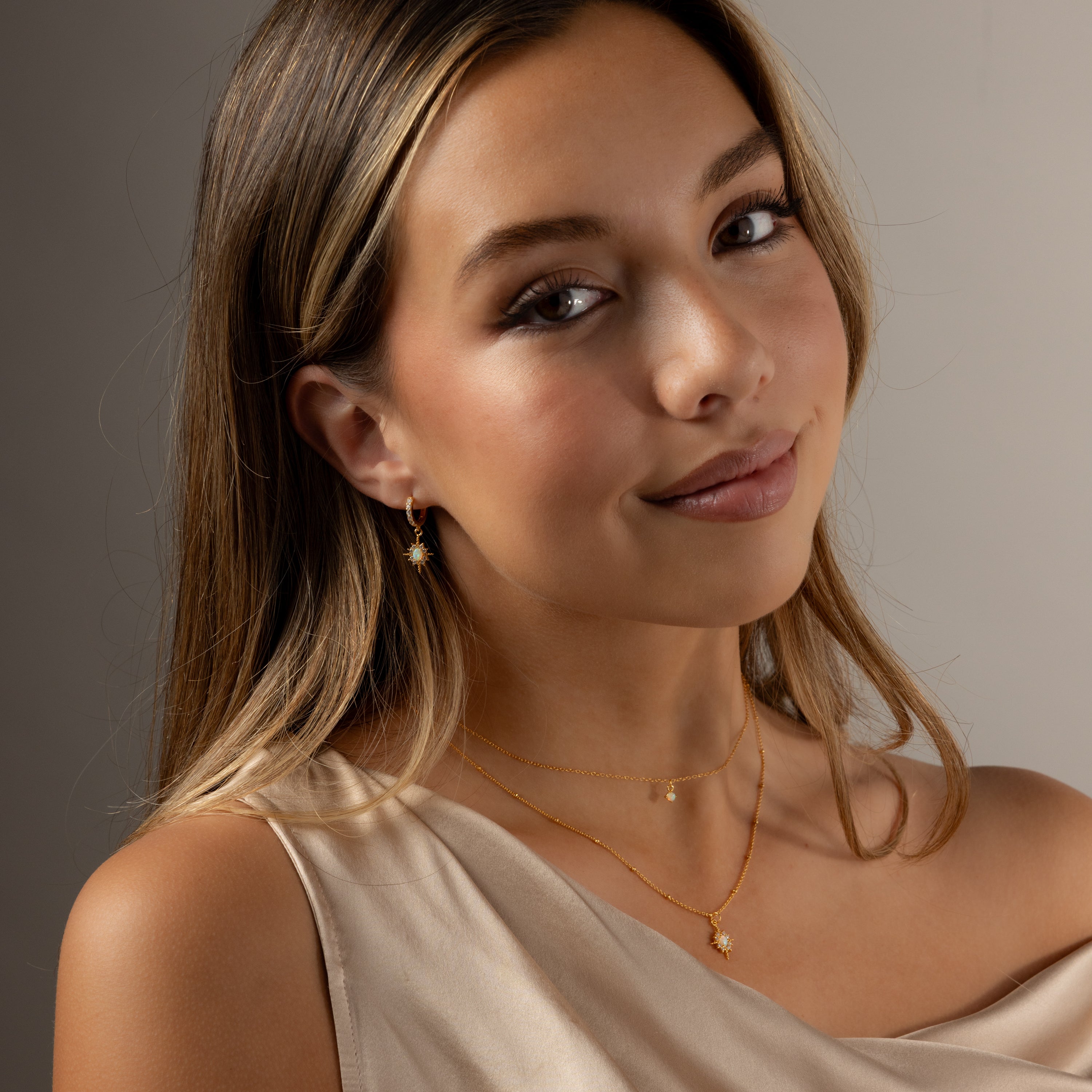 Young woman with long hair, wearing gold jewelry—Opal Star Necklace & Earrings Set—and a cream sleeveless top, smiles softly at the camera.
