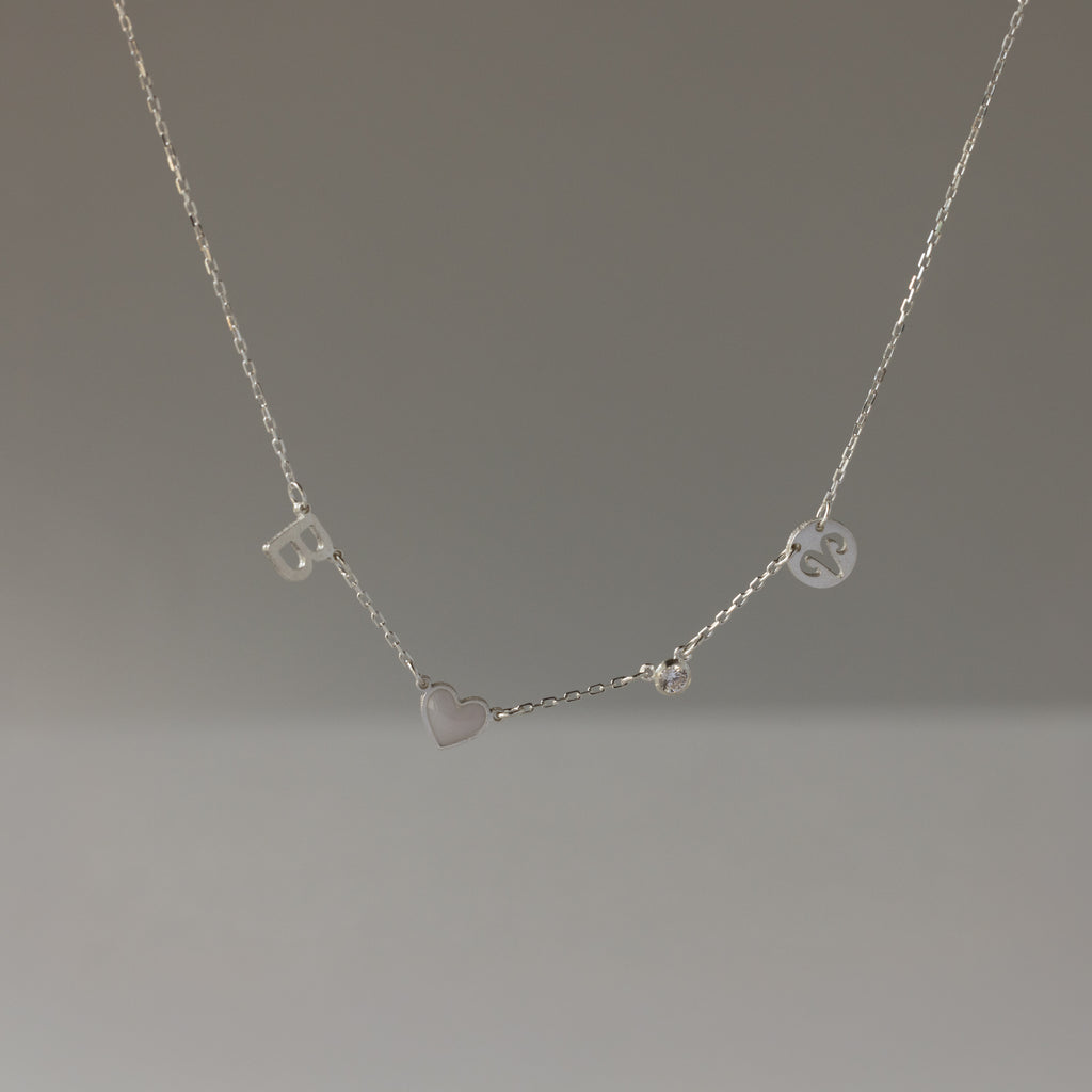 The Dainty Space Charm Necklace in Sterling Silver features letter, heart, and circular charms on a thin chain—perfect for adding a birthstone and personal touch.