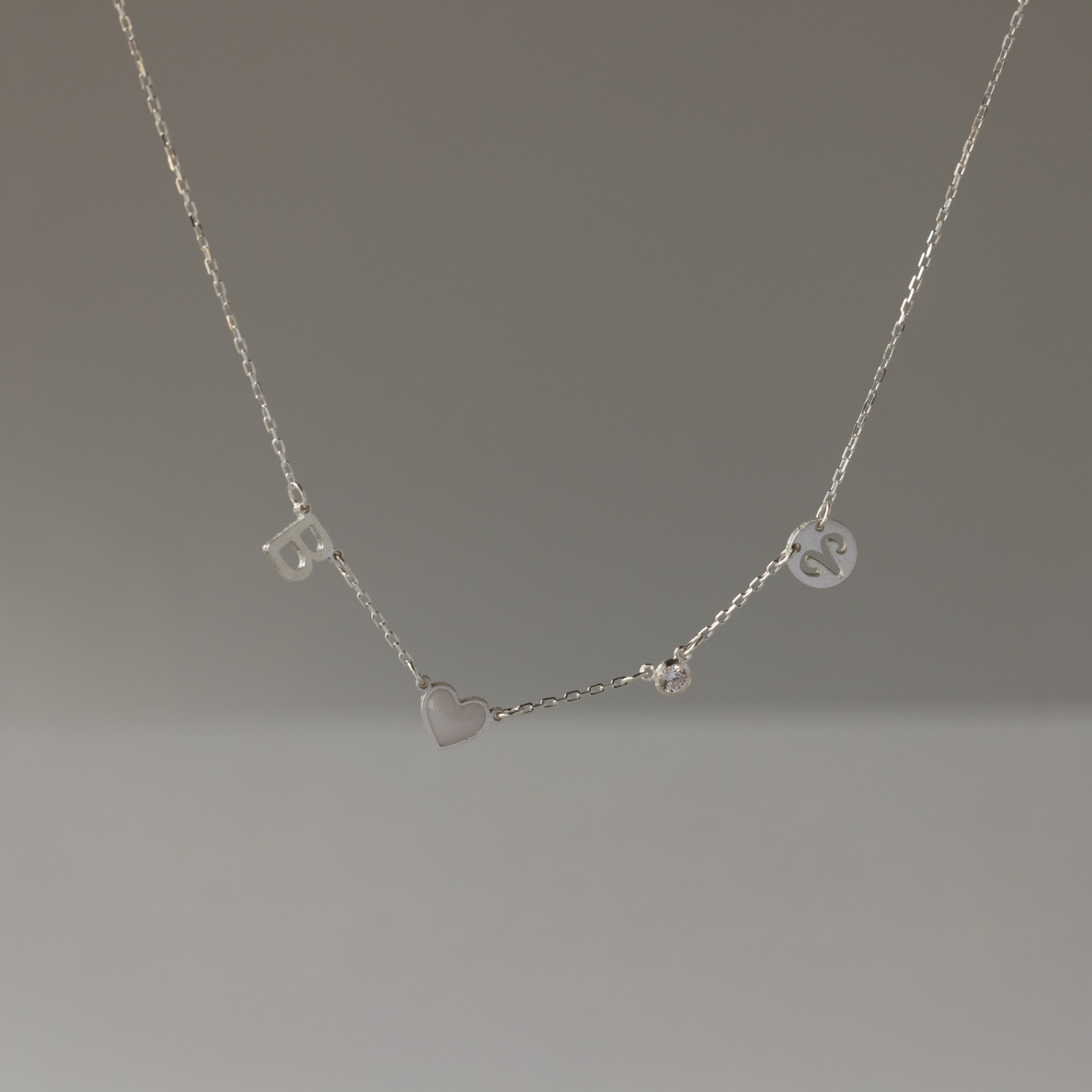 The Dainty Space Charm Necklace in Sterling Silver features letter, heart, and circular charms on a thin chain—perfect for adding a birthstone and personal touch.